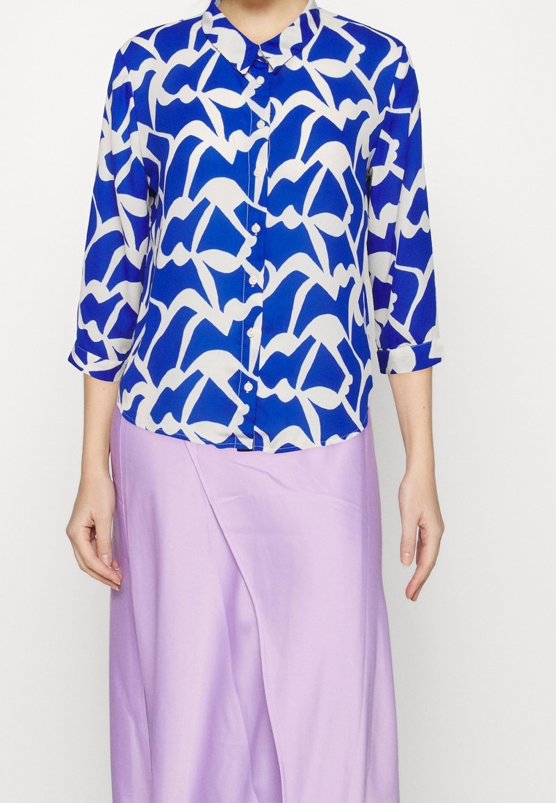 Blusa a righe blu e bianche con colletto, maniche di tre quarti e bottoni davanti. Indossata sopra una gonna lilla chiaro.