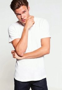 Bílý bavlněný tričko s krátkými rukávy, kulatým výstřihem a přilnavým střihem, spojené s tmavými džíny, na světlém pozadí.