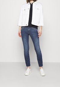 Vit denimjacka över en svart skjorta, i kombination med blå skinny jeans och vita sneakers. Funktioner inkluderar knappar, fickor och upprullade ärmar.