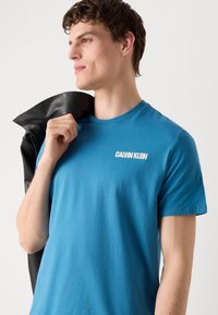 T-shirt in cotone blu con scollo rotondo e maniche corte, con il logo bianco "CALVIN KLEIN" sul petto. Il modello tiene in mano una giacca nera.
