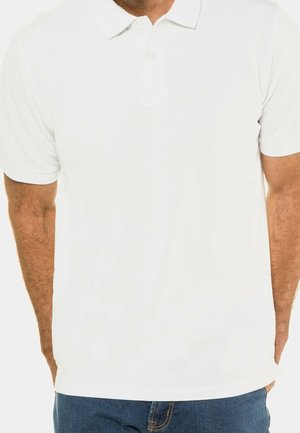 Mand iført en simpel hvid kortærmet polo og blå jeans, stående med hænderne i lommerne mod en hvid baggrund.