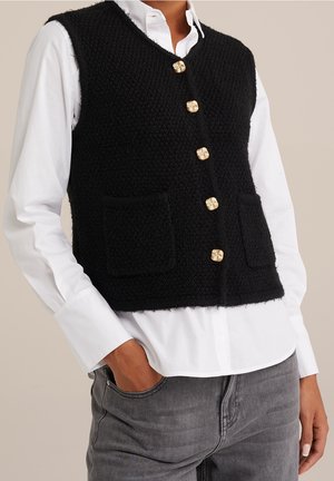 Veste sans manches noire texturée avec cinq boutons dorés en spirale portée sur une chemise blanche et un jean gris, une main dans la poche.