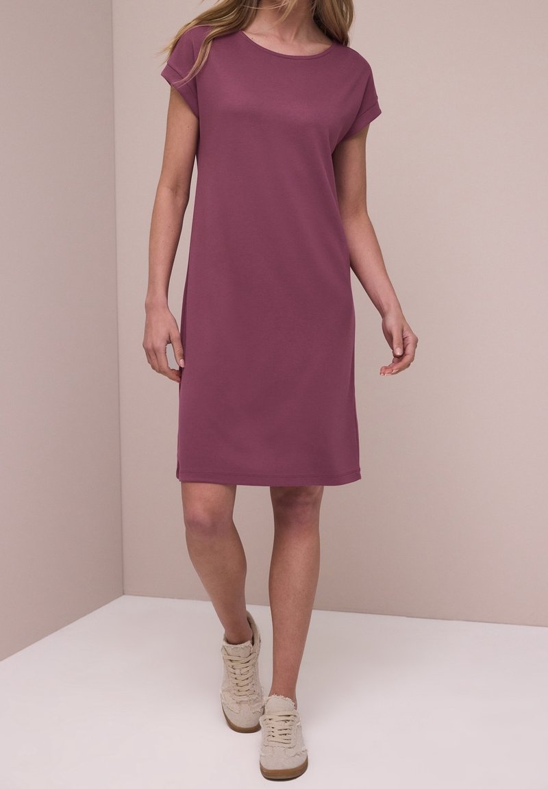 Femme portant une robe violette simple à manches courtes, longueur genou, et des baskets beiges à lacets, debout devant un fond beige uni.