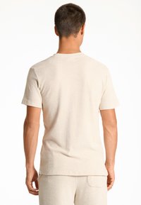 Camiseta de manga corta color beige claro, textura suave, cuello redondo, diseño simple, corte ajustado. Lleva puesta una persona con el cabello corto, vista desde atrás.