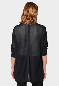 Mörk, löst sittande tröja med en texturerad stickad mönster, långa ärmar och en slitsdetalj bak. Materialet verkar lätt och semitransparent.