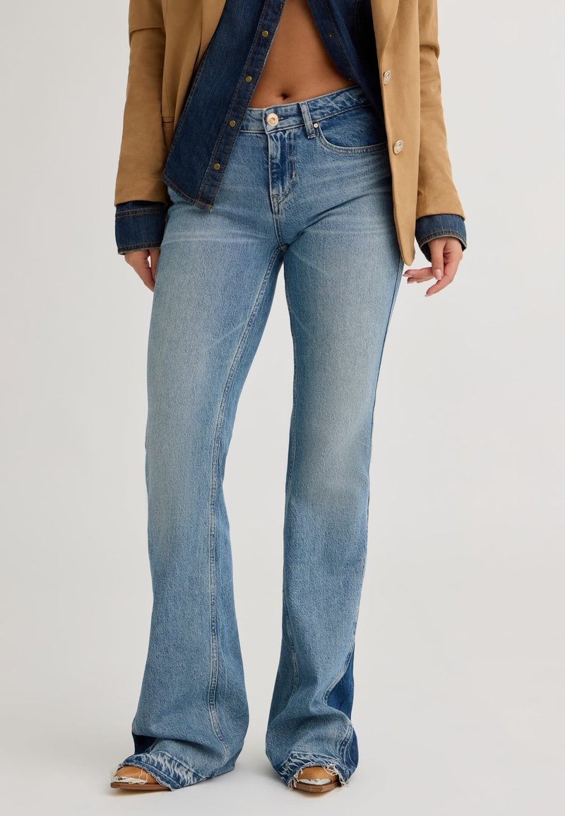 Jean évasé bleu clair taille mi-haute avec ourlets bruts et effilochés, associé à un blazer beige porté sur une chemise en denim foncé laissant apparaître le ventre.
