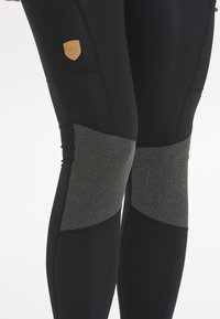 Musta leggingit, joissa on teksturoitu musta ja harmaa polven paneeli, ja joissa on pieni vaaleanruskea logo-ompelus. Farkkumainen kangas tiukalla istuvuudella ja ompeluyksityiskohdilla.