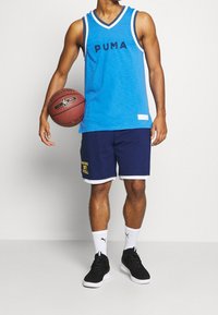 Man i blå Puma linnen och marinblå shorts som håller i en Spalding NBA basketboll, iklädd vita Puma-strumpor och svarta träningsskor.