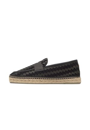 Zapato negro tejido tipo slip-on con suela trenzada de yute, tacón plano y una correa lisa sobre la parte superior, mostrado de perfil sobre un fondo blanco.