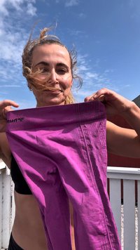 Vrouw buiten houdt paars leggings met beide handen omhoog tegen een blauwe lucht, haar blaast iets in de wind.