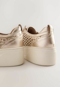 Metaliczne złote sneakersy z plecionym wzorem, okrągłym noskiem i białą platformą. Gładka tekstura z detalami przeszycia na brzegach.