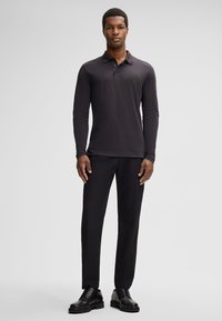 Langärmliges, dunkelgraues Poloshirt aus weichem Stoff, kombiniert mit schwarzen Hosen und schwarzen Schuhen. Das Modell steht vor einem neutralen Hintergrund.
