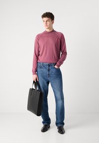 Dolga rokavna maroon majica z rebrastim ovratnikom, modri jeans in črna usnjena torba. Model stoji bos proti enostavni ozadju.