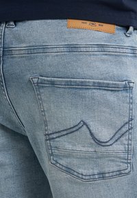 Vybledlé světle modré džíny z denimu s koženou štítkem v pase a jednou zadní kapsou s designem zakřiveného stehu.