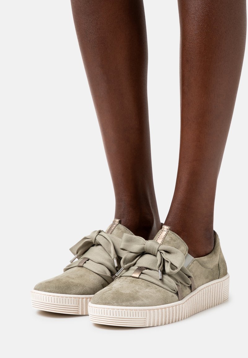 Jasnozielone zamszowe sneakersy z grubymi białymi gumowymi podeszwami i dużymi materiałowymi sznurowadłami związanymi na kokardki na stopach modelki.
