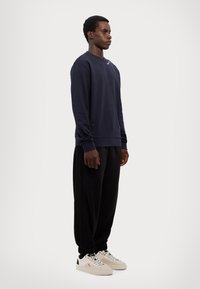 Granatowy sweter crewneck, czarne spodnie z plisami i kremowe sneakersy. Sweter ma prążkowane mankiety; spodnie mają elastyczne mankiety i luźny krój.