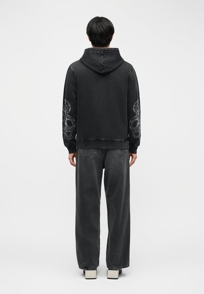 Άτομο με την πλάτη στραμμένη, φοράει ξεθωριασμένο μαύρο φούτερ με κουκούλα με σχέδια φιδιών στα μανίκια, φαρδιά σκουρόχρωμα τζιν και ανοιχτόχρωμα παπούτσια σε λευκό φόντο.