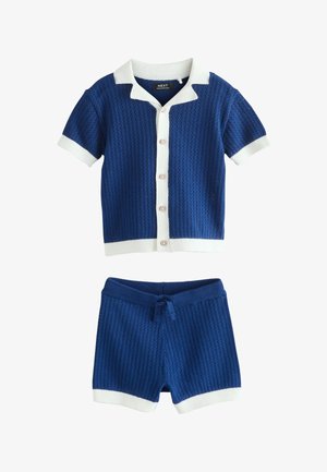 Plava vesteća košulja kratkih rukava s bijelim ovratnikom i manšetama, u kombinaciji s pripadajućim plavim vestećim kratkim hlačama s bijelim obrubom i strukom na vezanje.