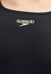 Μαύρο μαγιό από ύφασμα με κίτρινο λογότυπο Speedo, τοποθετημένο στο κέντρο κάτω από το ντεκολτέ στο άνω μέρος του στήθους ενός ατόμου.