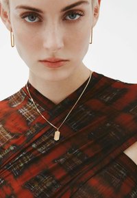 La modelo lleva una prenda de color rojo oscuro con un patrón y un leve brillo, con un escote en V. Se pueden ver pendientes de aro dorados y un collar delicado con un colgante.