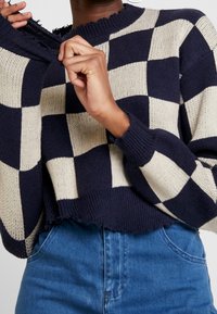 Pull en maille écossaise bleu marine et crème avec un col ras du cou côtelé et un ourlet effiloché, porté avec un jean en denim taille haute.