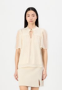 Krembluse med pufferm, prikkete tekstur, volangkrage og knytelukking. Brukt sammen med et beige kordfløyelskjørt med sidelåsing.