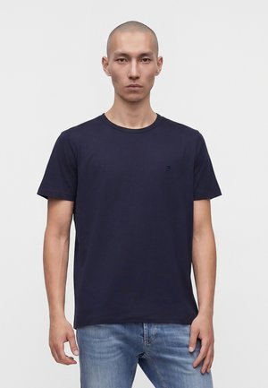 Jeune homme à la tête rasée portant un t-shirt bleu marine uni et un jean bleu clair sur fond blanc uni.