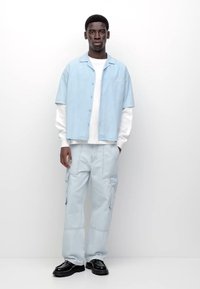 Camicia azzurra a maniche corte con bottoni sopra una camicia bianca a maniche lunghe, abbinata a pantaloni cargo azzurri e scarpe nere. Design semplice, vestibilità casual.