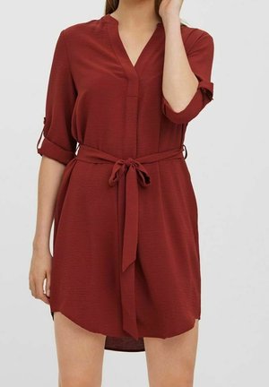 Kvinde iført en rustrød kjole med bælte og opkrogede trekvartærmer mod en ensartet lys baggrund.