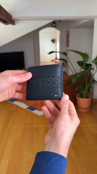 Svart läderplånbok med texturerat band, dubbelvikt design och präglad logotyp. Blanka metallaccenter, med en slät yta och kompakt form.