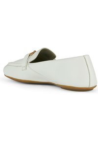 Zapato slip-on de cuero blanco con perfil bajo, costuras delineadas y suela de goma flexible; cuenta con una pequeña correa decorativa y un acento dorado.