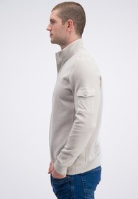 Beige pullover met een hoge kraag, gestructureerde brei-details op de mouwen en een zak op de linkerarm. Slim fit met geribbelde boorden en zoom.