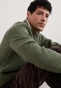 Pullover verde in maglia con una texture a coste, collo a giro, e dettagli variegati, abbinato a pantaloni di velluto a coste marroni. Design semplice e casual.