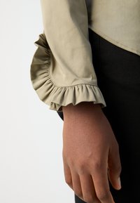 Blusa bege clara com punho frisado, textura suave, forma ajustada e costura em contraste, usada sobre calças pretas de tecido.