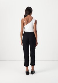 Femme aux longs cheveux noirs portant un haut blanc à une épaule, un pantalon noir court et des chaussures noires, debout devant un fond blanc uni.