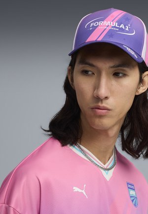 Młody mężczyzna z ciemnymi włosami do ramion, ubrany w różową koszulkę wyścigową Puma i fioletowo-różową czapkę Formula 1 Racing League.