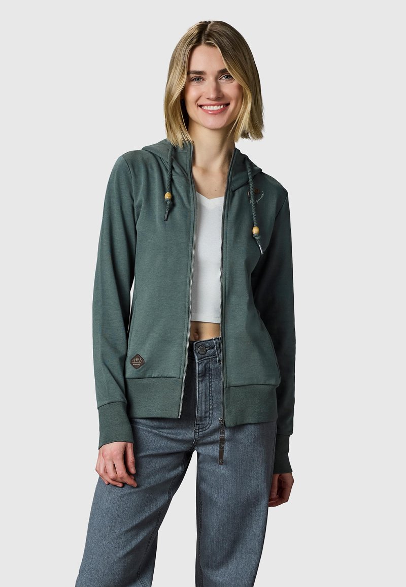 Jeune femme souriante, portant un sweat à capuche zippé vert foncé avec cordons et un jean bleu clair, se tenant devant un fond gris clair uni.