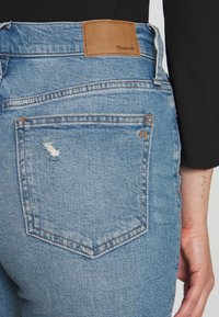 Jeans de denim azul claro com um bolso traseiro desgastado, etiqueta de couro beje na cintura e costura detalhada.