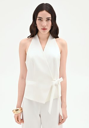 Donna con capelli scuri e mossi che indossa un gilet bianco senza maniche annodato in vita, pantaloni bianchi e chunky braccialetti dorati, rivolta in avanti.