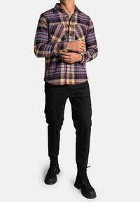 Koszula flanelowa w kratę w odcieniach niebieskiego, czerwonego i beżowego, z dwoma kieszeniami na piersiach i kołnierzykiem w kształcie ostrym, w połączeniu z czarnymi spodniami cargo i butami.