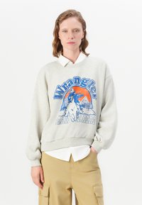 Šedá mikina s modrou grafikou "Wrangler Trail Riders", balónové rukávy a ribovaný lem cez bielu košeľu s golierom. Neformálny dizajn.