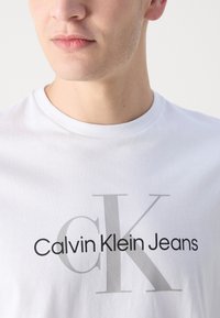 T-shirt in cotone bianco con la scritta "Calvin Klein Jeans" in caratteri neri e grigi, collezione a girocollo e tessuto morbido.