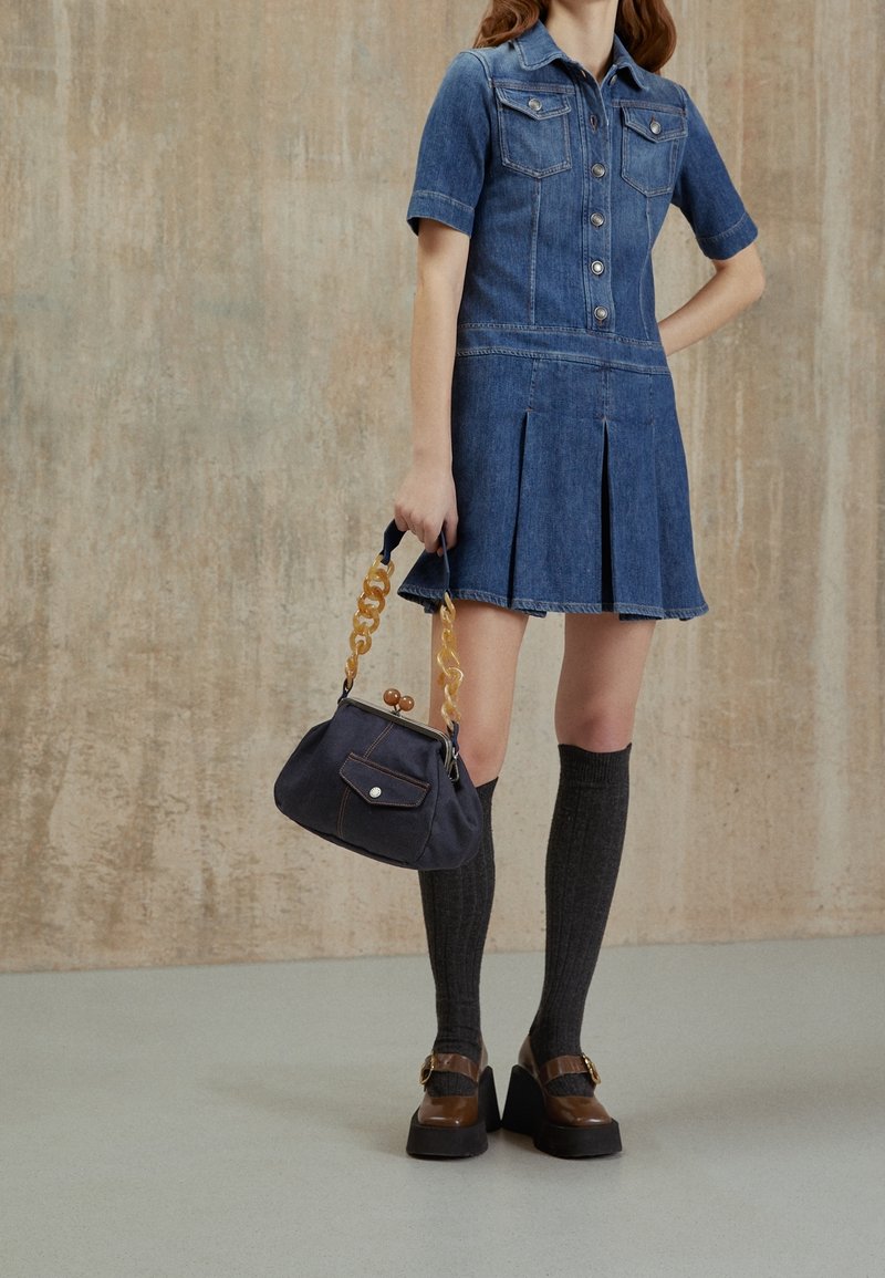 Vestido de mezclilla con mangas cortas, bolsillos frontales y falda plisada; combinado con un bolso oscuro que presenta una correa de cadena con patrón y acentos dorados.