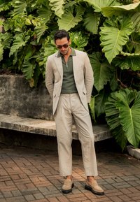 Traje de lino beige claro con pantalones ajustados, combinado con una camiseta tipo polo verde con cremallera y mocasines beige. Rodeado de grandes hojas verdes.