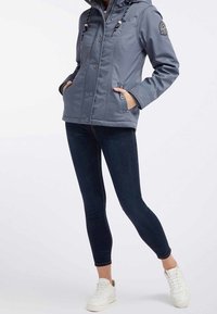 Ung vuxen iklädd en grå huvtröja, mörka jeans och vita sneakers, stående framför en neutral bakgrund och visar upp en avslappnad outfit.