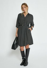 Schwarzes Polka-Dot-Kleid mit Kragen, Dreiviertelärmeln, tailliertem Gürtel, schwingendem Rock, kombiniert mit kniehohen schwarzen Lederstiefeln und einer Tasche.