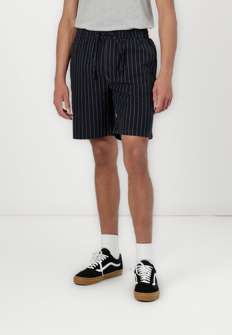Shorts rayés bleu marine avec une taille élastique et un cordon de serrage. Associés à des baskets noires et blanches et des chaussettes blanches mi-mollet.