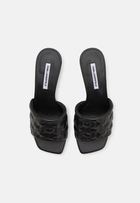 Chanclas negras con una correa texturizada y acolchada y suela lisa. Presenta una etiqueta con el logo en la plantilla, diseño minimalista.