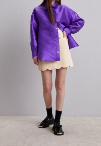 Chemise en satin violet à boutons, jupe beige à ourlet festonné, chaussures noires à pampilles, associées à des chaussettes noires. Design simple et moderne.