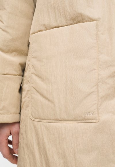 Μανίκι μπεζ μπουφάν με επένδυση και μεγάλη μπροστινή τσέπη που φέρει διακριτική κέντηση "MM" κοντά στην κάτω δεξιά γωνία.